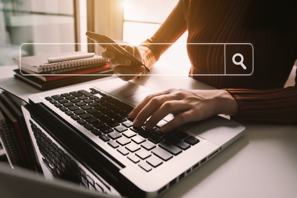 AdobeStock 279957470 Personne tapant sur un ordinateur avec un smartphone à la main, symbolisant une recherche internet